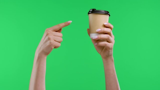 A woman's hand holds a paper cup with coffee, the other hand points with a forefinger to coffee and shows a thumb up. Close up of coffee cup and woman hands on green screen in studio. Slow motion.