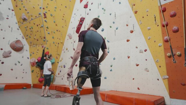 Rear-view low-angle medium slowmo of male athlete with leg prosthesis standing at modern indoor sports complex watching people climbing wall