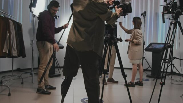 Tilt-up rear-view slowmo of professional cameraman with robotic leg walking into filmmaking studio and starting setting up camera and getting ready for video shooting. Crew helping him