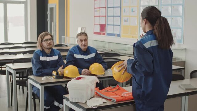 Medium slowmo of female factory worker explaining safety rules for manufacturing facility safety to male coworkers listening to her while sitting at desk in class holding yellow hard hats