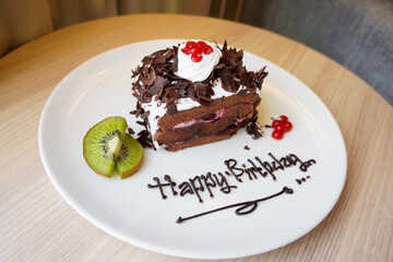 Close up top view of chocolate birthday cake with whipped cream, sliced kiwi and cherry on white plate at the table. Bokeh background. No people.
