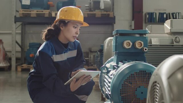 Medium slowmo of female plant worker in hard hat and uniform squatting next to machinery equipment operating it with digital tablet in hands