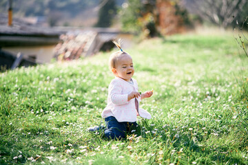 Laughing little girl with a ponytail on her head sits on her knees on a green lawn among white...