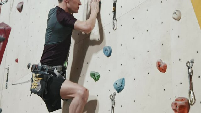 Tracking shot of strong athletic man with amputated leg climbing wall indoors at modern sports centre wearing safety equipment. Concept of sports motivation for disabled people