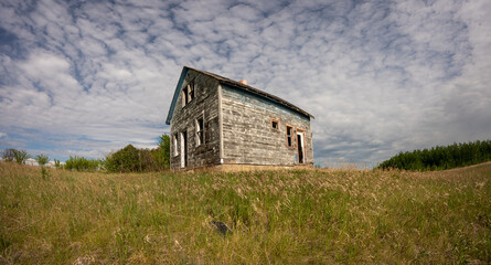 Obraz premium Low angle perspective of a dilapidated wooden farmhouse with long grass and missing windows. The sky is blue with white clouds. 