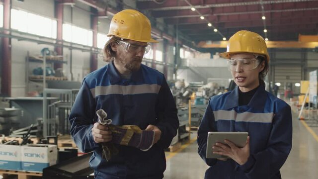 Medium slowmo of couple of professional factory workers discussing project plan looking at tablet while walking through plant facility wearing safety glasses and hard hats
