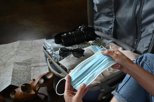 Close-up of woman's hands holding surgical masks, suitcase with clothes, sunglasses, photo camera, map, sandals, copy space