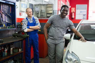 Smiling satisfied male client standing touching his repaired car in auto repair shop