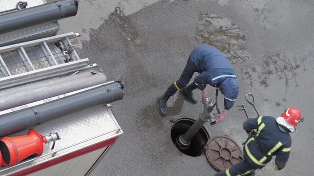 Man in jumpsuit unscrews hydrant from rusty sewer hatch under heavy rain near fire car slow motion upper view. Concept firefighters training