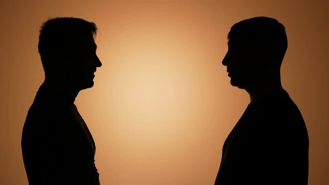 Two men talk, chat, clink beer bottles and drink beer, laugh, have fun. Silhouettes of two men in the studio on a brown gradient background. Meeting friends enjoying drinks. Slow motion. Close up.