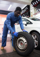 Fototapeta na wymiar Afro american auto mechanic in uniform holds wheel at tire service
