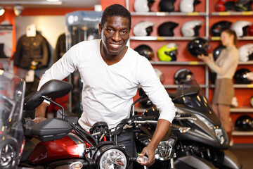 Fototapeta na wymiar Lucky Afro American man posing near new purchased motorcycle in bike salon