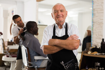 Obraz premium Portrait of professional aged barber standing with crossed arms in modern hair salon..