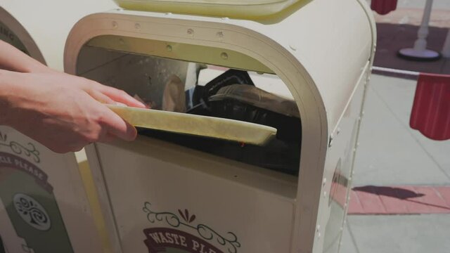 This slow motion video shows anonymous customer hands cleaning their fast food tray off and throwing away the trash from their meal into an outdoor, public trash receptacle.