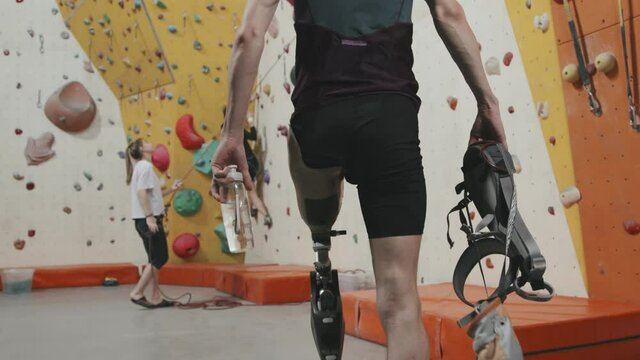 Medium slowmo of sportsman with leg prosthesis holding safety equipment for wall climbing while walking along sports complex then sitting on bench to take prosthesis off