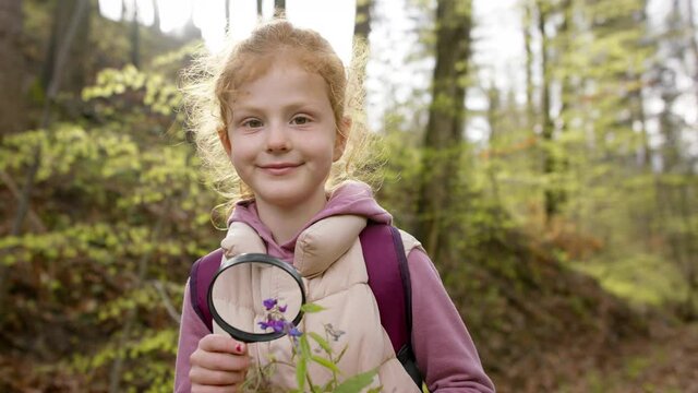Small girl examining flower with magnifying glass in nature, looking at camera.