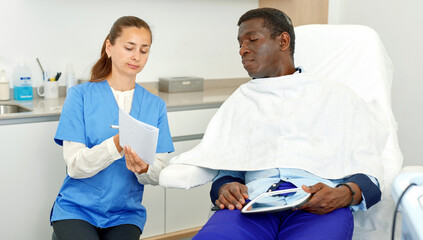 Professional cosmetician woman with papers talking to man before procedure in clinic of esthetic cosmetology