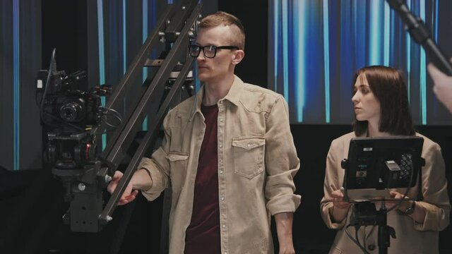 Medium shot of professional cameraman shooting video in studio using crane while his female assistant helping him with storyboard and gaffer holding lighting