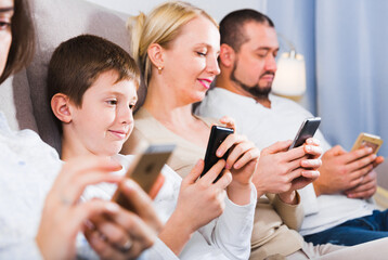 Young parents with son using mobile gadgets on sofa at home