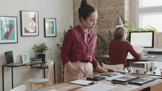 Medium PAN slowmo of female architect choosing building material for new house looking at its 3d layout on work desk while her colleague working on computer in background