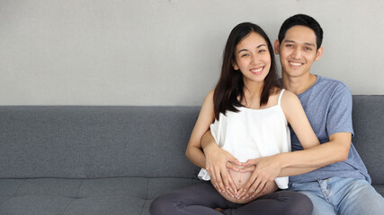 Young asian husband embraces young asian pregnant wife and keep hand on her belly, enjoying become parents soon  while they doing heart symbol. They sitting on sofa and look happy and love together