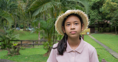 Portrait happy asian young girl wearing straw hat staying in forest park. Female youth volunteer guide standing on the lawn to welcome tourists in tropical forest. - 436967519