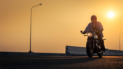 A man biker in a denim jacket drives a vintage motorcycle on a highway at sunset. Man on Vintage Bike. Biker Lifestyle Concept.
