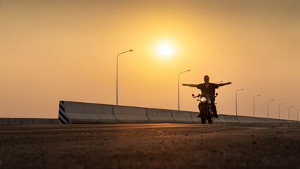 Fototapeta na wymiar A man riding a vintage motorcycle with spread arms on the highway at sunset. Man on Vintage Bike. Biker Lifestyle Concept.