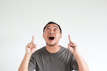 Wow and surprised face expression of Asian man in grey t-shirt with hand pointing up on empty space