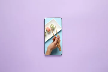  Female hand with chopsticks and sushi rolls on screen of mobile phone on color background © Pixel-Shot