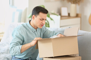 Upset young man unpacking parcel at home - obrazy, fototapety, plakaty