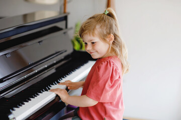 Beautiful little toddler girl playing piano in living room. Cute preschool child having fun with learning to play music instrument. Early musical education for children.
