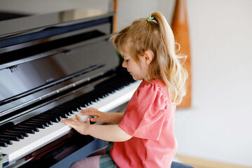 Beautiful little toddler girl playing piano in living room. Cute preschool child having fun with learning to play music instrument. Early musical education for children.
