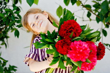 Obraz na płótnie Canvas Cute adorable little toddler girl with huge bouquet of blossoming red and pink peony flowers. Portrait of smiling preschool child in domestic garden on warm spring or summer day. Summertime.