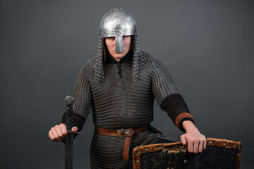 Portrait of a medieval warrior of the late viking era and the beginning of the crusades sitting on his knee. Knight in chain mail and helmet armed with shield and sword isolated on dark background.