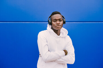 Young African American black man listening music with wireless headphones while wearing a white sweatshirt, looking to camera with serious expression and arms crossed