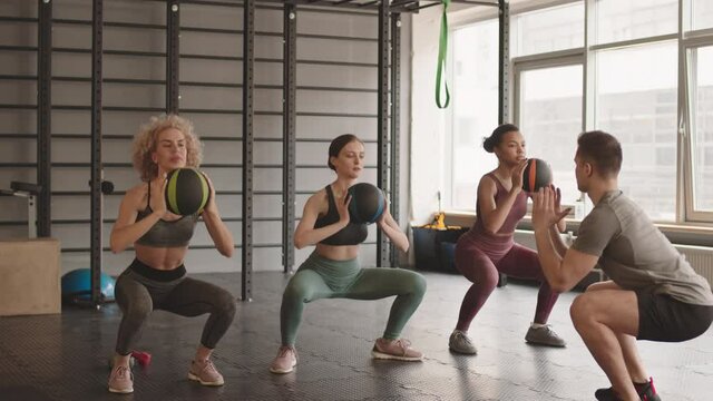 Wide shot of three slim Caucasian and Mixed-race women wearing sportswear, doing squats holding balls in hands in front of them, looking at male fitness coach