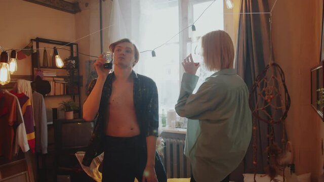 Arc shot of young tipsy couple dancing in bedroom decorated with garland, doing love shot and drinking alcohol at home party