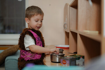cute caucasian boy in carnival costume of bear playing with toy kitchen in kindergarden pretending...