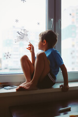 portrait of caucasian 6-7 years old boysitting on sill and  dreaming about christmas touching a deer sticker on wind. Staying home because cold. Image with selective focus and noise effect