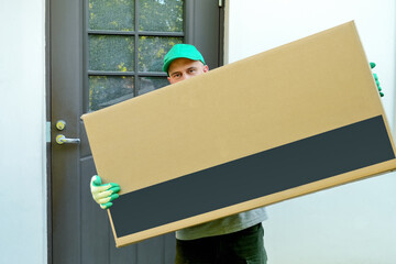 Delivery man holding a large cardboard box at the door of the house. The postal courier delivered the parcel to the address. Works on transportation and delivery of parcels. Space for text and design.