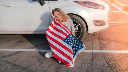 Young millennial blonde woman sitting with american flag next to car. Flag of the united states in her hands. July 4th Independence Day. USA national holiday. Freedom and memorial concept