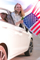 Young millennial blonde woman looking from car with american flag. Flag of the united states in her...