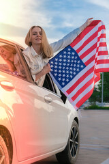 Young millennial blonde woman looking from car with american flag. Flag of the united states in her...