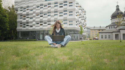 Fototapeta na wymiar Busy attractive girl working at the laptop as sitting on grass in city park on hectic summer morning, outdoor shot in urban area. Student studying on computer sitting on campus staircase outside.