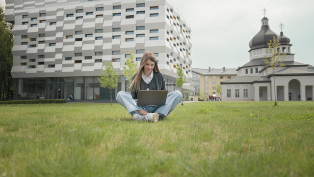 Busy Attractive Girl Working At The Laptop As Sitting On Grass In City Park On Hectic Summer Morning, Outdoor Shot In Urban Area. Student Studying On Computer Sitting On Campus Staircase Outside.