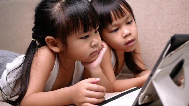 Two sibling girls lying on the sofa and watching digital tablets together in the living room.