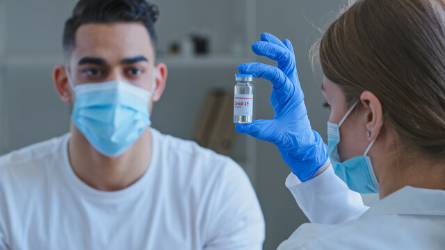 View From Behind Caucasian Unrecognizable Woman Doctor Wears Medical Gloves Holds Jar With Covid-19 Virus Vaccine Drug Shows Arabic Spaniard Guy Man Patient In Face Mask Sitting At Table, Vaccination