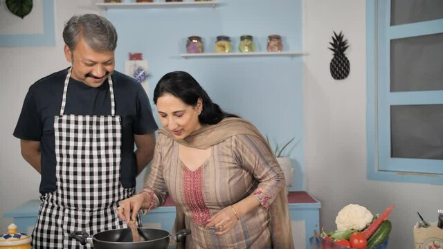 Happy male watching his wife tasting food from the pan - cooking concept. Medium shot of an attractive mature couple making a dish in their kitchen - caring husband / home-style cooking