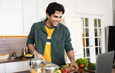 Fototapeta na wymiar Young smiling man cooking food while looking at online recipe while standing at home kitchen